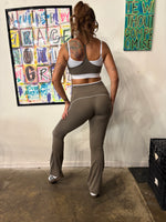 Load image into Gallery viewer, A woman stands on a runway wearing a matching beige athletic set consisting of a sports bra and flared leggings with white contrast trim. 
