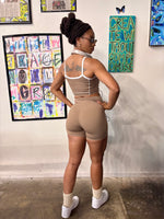 Load image into Gallery viewer, A woman stands with her back to the camera wearing a beige athletic set consisting of a sports bra and fitted shorts, 

