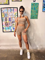 Load image into Gallery viewer, A woman stands in a studio wearing a beige two-piece athletic set with white contrast stitching, consisting of a crop top and matching shorts. She is accessorized with white sneakers, white socks, and over-ear headphones around her neck
