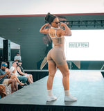 Load image into Gallery viewer, A woman models a beige two-piece athletic set consisting of a tank top and matching shorts, standing on a stage. The set is light tan/beige with white trim.
