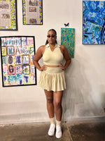 Load image into Gallery viewer, A woman modeling a cream-colored two-piece ribbed set consisting of a halter crop top and a pleated skort 
