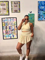 Load image into Gallery viewer, A woman posing in a matching cream-colored two-piece activewear set consisting of a halter crop top and a pleated mini skort
