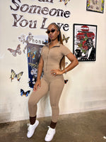Load image into Gallery viewer, A woman models a beige short-sleeve zip-up one-piece jumpsuit, styled with white sneakers, brown socks, and sunglasses
