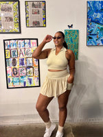 Load image into Gallery viewer, A woman posing in a cream-colored two-piece athletic set consisting of a sleeveless crop top and a pleated mini skort
