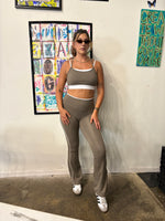 Load image into Gallery viewer, A woman stands on a runway wearing a matching beige athletic set consisting of a sports bra and flared leggings with white contrast trim. 

