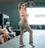 Load image into Gallery viewer, A woman stands on a runway wearing a matching beige athletic set consisting of a sports bra and flared leggings with white contrast trim. 
